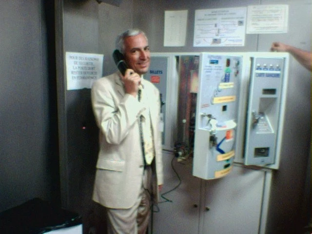 Un homme en costume blanc parle au téléphone dans une cabine téléphonique. Derrière lui, des panneaux et des équipement